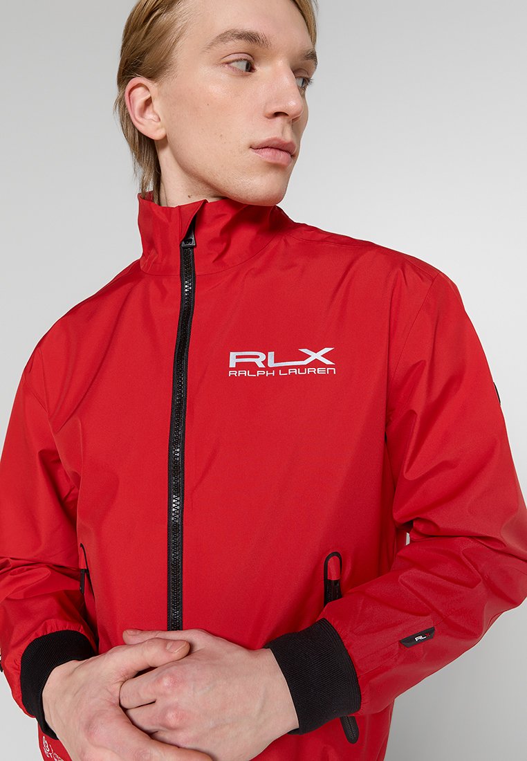 Chaqueta impermeable roja con cuello alto, cierre de cremallera, acentos negros y logo de RLX. Las características incluyen puños elásticos y bolsillos frontales.