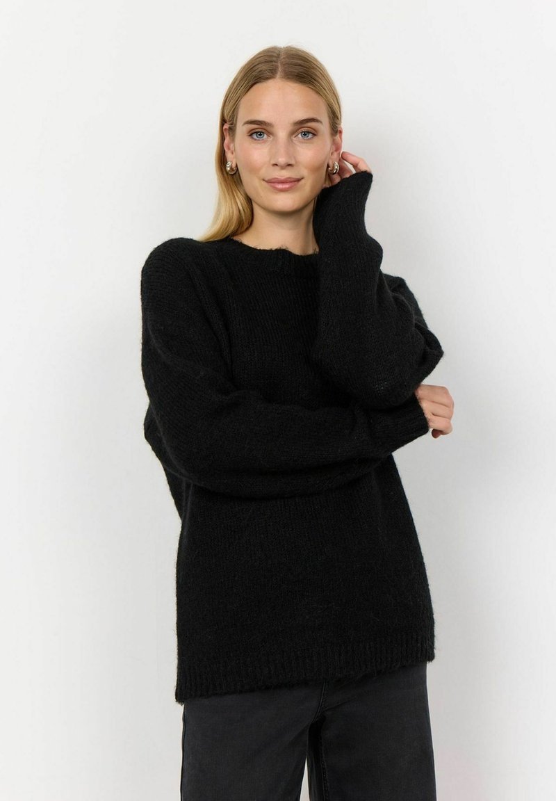Pullover negru supradimensionat cu o textură cu dungi, mâneci largi și un tiv brut. Stilizat cu pantaloni negri cu talie înaltă pe un fundal simplu.