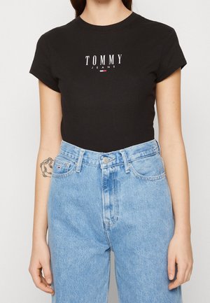 Person trägt ein schwarzes Tommy Jeans T-Shirt und hoch taillierte hellblaue Jeans, mit einem Kettentätowierung am linken Unterarm.