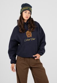 Granatowy sweatshirt z dużym kapturem, złotym haftem herbu i napisem "Social Club", w połączeniu z brązowymi spodniami z zakładkami i pasującą czapką.