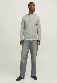 Man wearing light gray hoodie, faded blue jeans, and dark blue sneakers, standing and looking to his right against plain background.