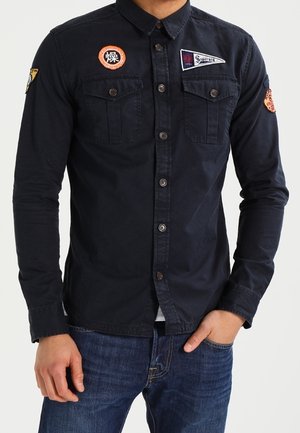 Mężczyzna ubrany w dopasowaną czarną koszulę zapinaną na guziki z kieszeniami na piersi i naszywkami, w niebieskich dżinsach, stoi z jedną ręką w kieszeni.