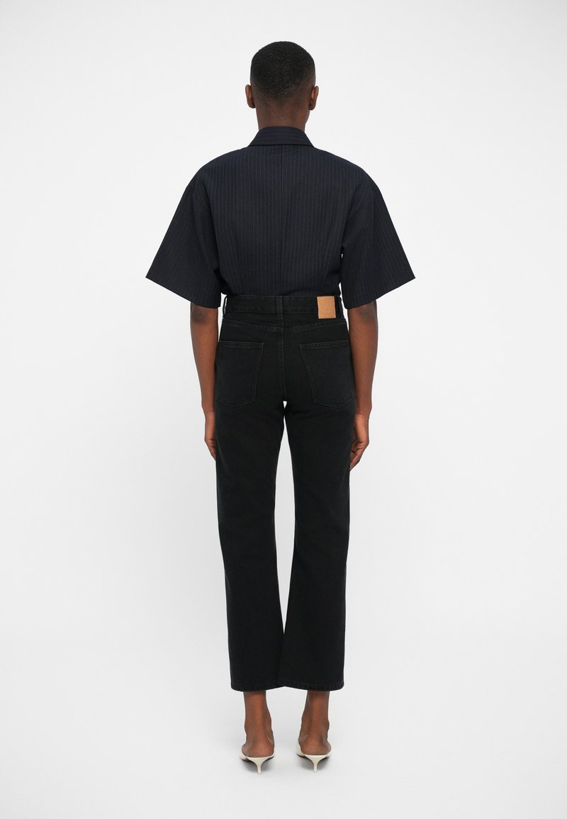 Pantalons en denim noirs avec une jambe droite et des poches au dos, associés à une chemise à manches courtes à rayures sombres. Design simple et épuré, avec une taille ajustée.