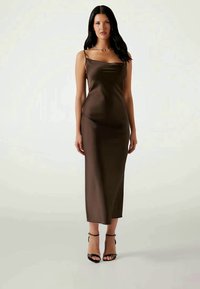 Woman with long dark hair wearing a sleeveless brown satin midi dress and black high-heeled sandals, standing against a plain background.
