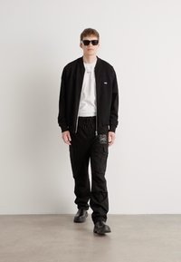 Schwarze Bomberjacke über einem weißen T-Shirt, kombiniert mit schwarzen Cargo-Hosen mit Taschen. Das Modell trägt schwarze Sonnenbrillen und Slipper.
