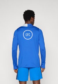 Blå langærmet sportstrøje med en lysere blå krave, der har et cirkulært "CFC" logo på ryggen. Sammen med blå shorts.