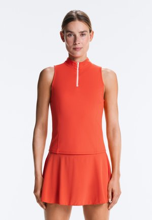 Žena v červených sportovních šatech bez rukávů se zipem vpředu stojící na jednobarevném světlém pozadí.