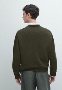 Tmavě olivově zelený svetr s kulatým výstřihem, vyrobený z texturované pleteniny. Má žebrované manžety a lem, spárovaný se světlými šedými kalhotami.