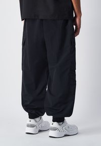 Zwarte cargobroek met zijzakken, elastische boorden en een gladde textuur, gecombineerd met witte sneakers met zwarte accenten.