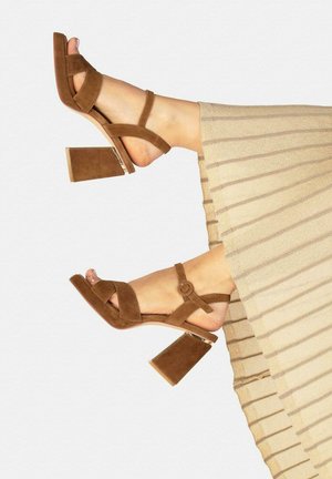 Pies con sandalias de tacón ancho de ante marrón, con tiras y hebillas, combinadas con pantalones plisados beige.