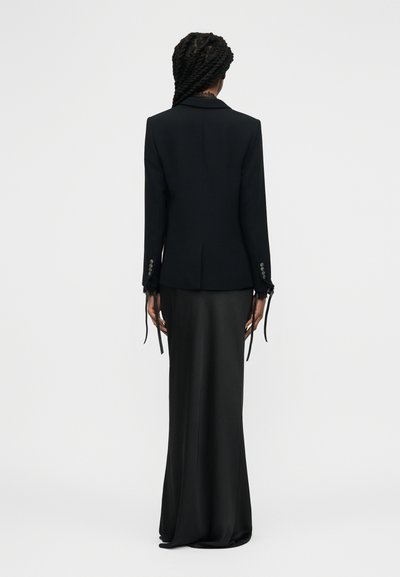 Femme aux longs cheveux tressés portant un blazer noir ajusté avec des poignets boutonnés et une longue jupe noire, debout de dos.