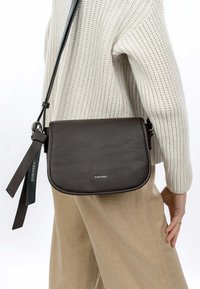 Borsa a tracolla in pelle marrone con texture perforata, marchio argentato e tracolla regolabile, indossata sopra un maglione chiaro e pantaloni beige.