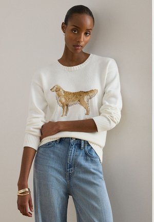 Ženska, oblečena v belo pulover z grafiko rjavega psa, oblečena v modre kavbojke z visokim pasom, stoji ob navadni svetli steni.