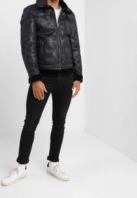 Giubbotto di pelle nera con profili in pelliccia sintetica, dotato di chiusura con zip e due tasche laterali. Indossato con jeans neri e scarpe da ginnastica bianche.