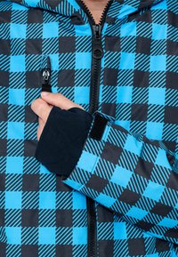 Chaqueta de cuadros azul y negro con un bolsillo con cremallera, tejido texturizado y puños elásticos, con patrones de acento de rayas verticales.