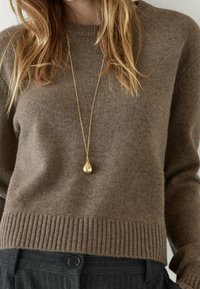 Maglione marrone lavorato a maglia con orlo a coste, abbinato a una collana con pendente a goccia dorato. Tessuto texturizzato, vestibilità comoda e design sobrio.