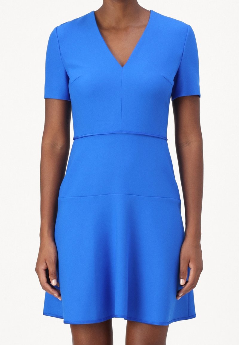 Femme portant une robe bleu roi à manches courtes, col en V, longueur genou, taille cintrée et jupe évasée sur fond blanc.