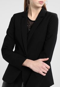 Blazer noir avec une coupe ajustée, revers crantés et une fermeture à un bouton. Texture de tissu lisse avec un effet superposé.