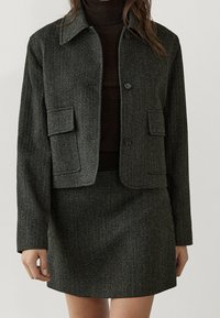 Szary cropped żakiet w desenie houndstooth z dwiema przednimi kieszeniami, w zestawie z pasującą spódnicą. Obie części mają teksturowaną tkaninę i zapięcia na guziki.