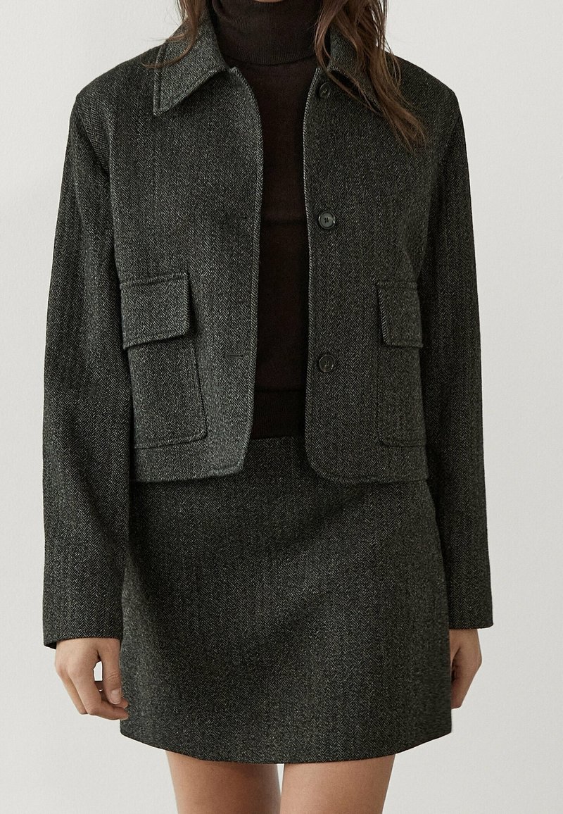 Szary cropped żakiet w desenie houndstooth z dwiema przednimi kieszeniami, w zestawie z pasującą spódnicą. Obie części mają teksturowaną tkaninę i zapięcia na guziki.