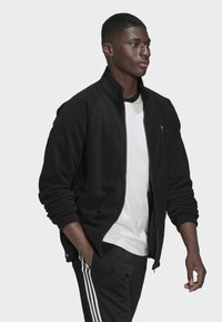 Must värvitud tõmblukuga fliisjakki, millel on soonikääred ja -kaelus. Kantud valge t-särgi kohal ja kombineeritud mustade pükstega, millel on valged triibud.