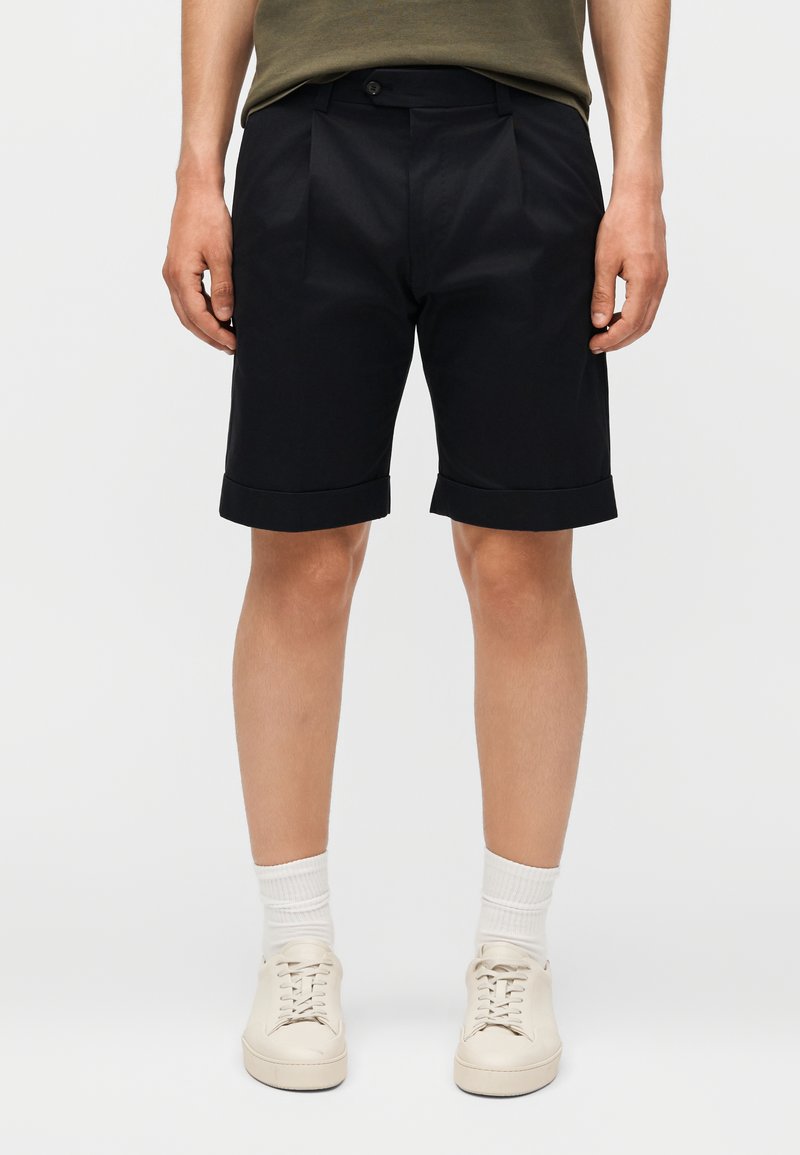 Άνδρας που φοράει μαύρα σορτς με στριφώματα, λευκές κάλτσες και λευκά αθλητικά παπούτσια, στέκεται μπροστά σε ένα απλό φωτεινό φόντο.