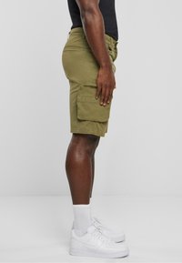 Pantaloni scurți cargo de culoare verde măslin, cu buzunare laterale mari, confecționați dintr-un material rezistent. Purtați cu adidași albi și șosete până la gleznă, evidențiind un profil lateral.