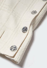 Tissu texturé crème avec un motif à carreaux subtil, décoré de quatre boutons ronds en argent avec des découpes fines et élaborées.