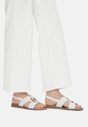 Sandalias planas blancas hechas de material suave, con una gran hebilla dorada y tiras cruzadas. La suela es plana y de color marrón claro.