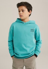 Turkos hoodie i bomull med en framficka med ett texturerat rutmönster och ribbade ärmar, bärs över beiga byxor.