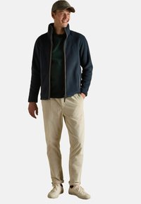 Tumesinine fliisjakk kergelt seistev laadiga krae, tõmblukk ees; rohelise särgi alla, kreemivärvilised sametpüksid ja valged tossud. Stilissecheckne kork.