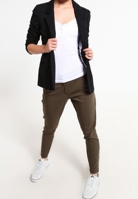 Černý blazer přes bílou košili s knoflíky, doplněný olivově zelenými zúženými kalhotami a bílými teniskami. Má upravený střih a hladké textury.