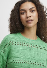 Pull en laine vert avec un col rond, présentant des rayures horizontales et de petites découpes sur l'ensemble du tissu.