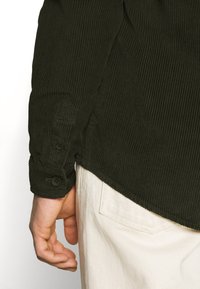 Dunkelgrünes geripptes Cordhemd mit Knopfmanschetten, kombiniert mit schmutzig-weißen Hosen. Der Stoff hat eine strukturierte Oberfläche.