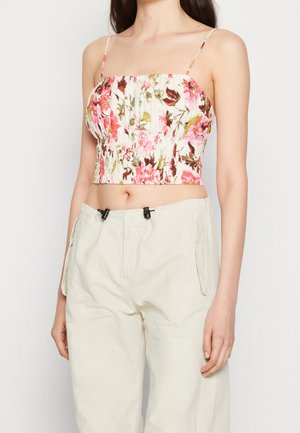 Kvinde iført en blomsterprikket pink og hvid crop top med tynde stropper og beige bukser med høj talje mod en ensfarvet baggrund.