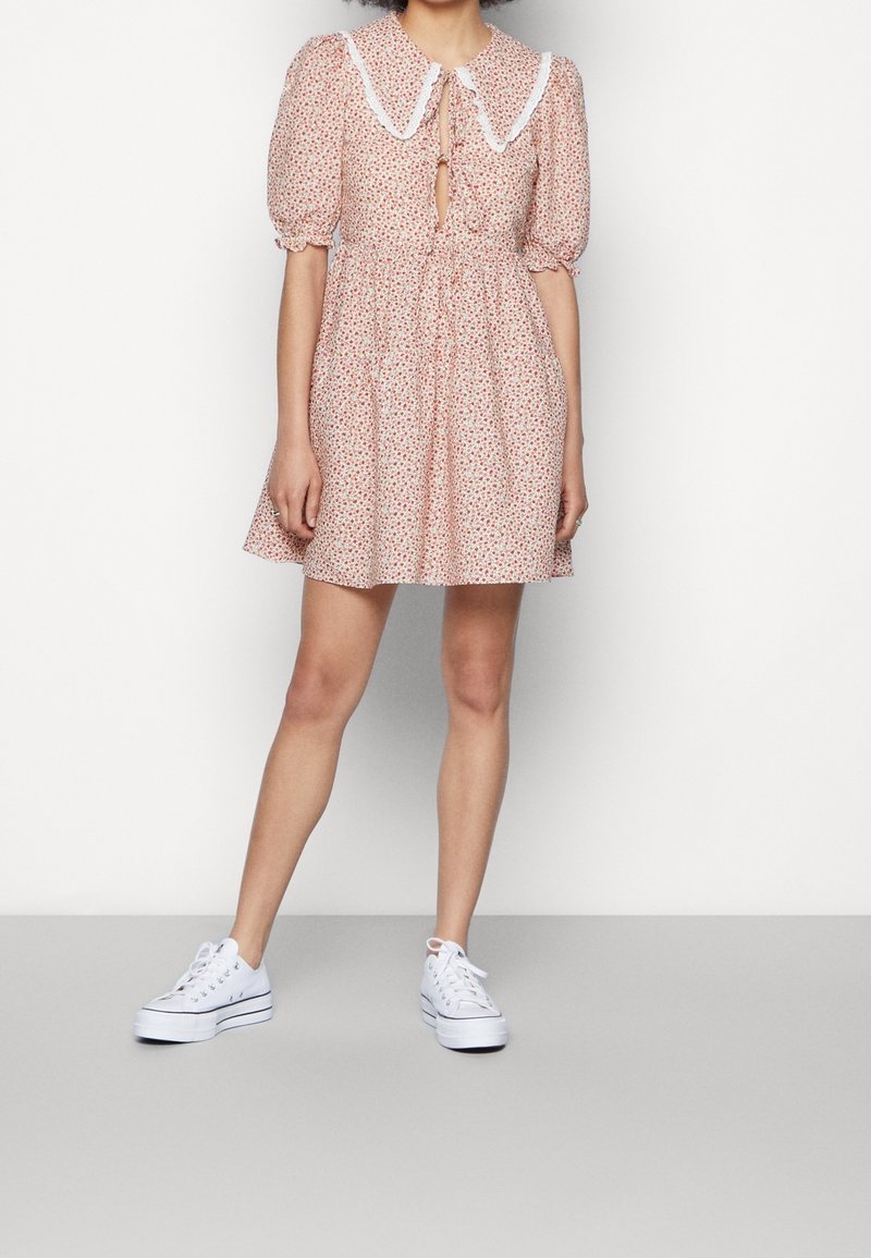 Femme portant une robe courte rose à fleurs avec des manches bouffantes, un large col orné de bordure blanche, et des baskets à plateforme blanches, debout contre un mur uni.
