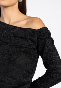 Zwarte off-shoulder top gemaakt van een gestructureerde, glinsterende stof. Kenmerkt lange mouwen met plooidetails en een aansluitend ontwerp.