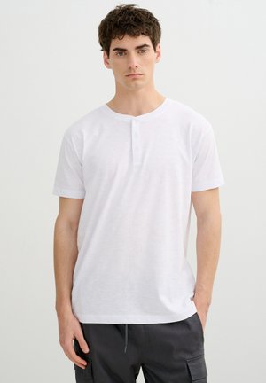 Joven con cabello oscuro y rizado, vistiendo una camiseta henley blanca de manga corta y pantalones cargo oscuros, de pie frente a un fondo claro y liso.