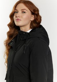 Cazadora negra impermeable con capucha, con cierre frontal de cremallera, cordón ajustable y bolsillos laterales. Textura suave con un sutil detalle del logo en la manga.
