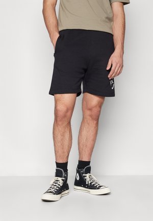 Mand iført sorte afslappede shorts med lommer, sorte højtop-sneakers og en beige t-shirt, stående mod en hvid baggrund.