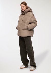 Giacca imbottita di colore marrone chiaro con colletto alto e cappuccio; presenta una forma squadrata e tasche a filetto; indossata con pantaloni verde scuro e sneakers bianche.
