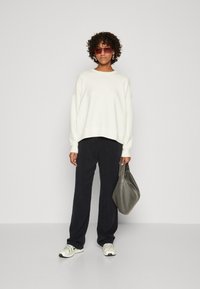 Lys krem oversize hettegenser, mørk marineblå vide bukser, grå veske og mønstrede sneakers. Krøllete hår og rosa solbriller.