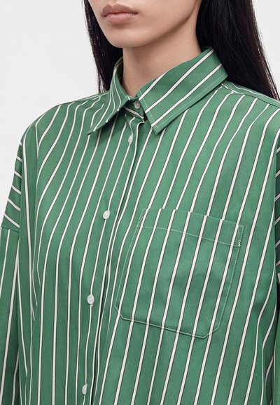 Femme portant une chemise verte à rayures blanches verticales avec une poche poitrine, montrée des épaules au menton.
