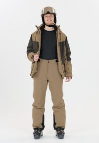 Skiausrüstung mit einer braunen Jacke und passenden Hosen, die eine strukturierte Oberfläche aufweisen, verstellbare Träger und schwarze Akzente haben. Getragen mit einem Helm und einer Skibrille.