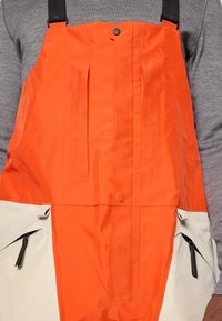 Kohvorage ja beežid põllülikonnad, millel on reguleeritavad mustad rihmad. Sisaldab voldikesi ja lukuga taskuid külgedel. Sile, tekstuuriga kangas.