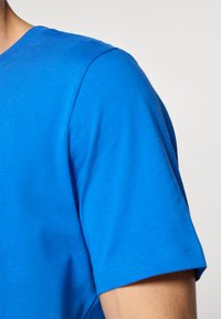 Tuva cilvēka, kurš valkā vienkāršu spilgti zilu īsroku T-kreklu, attēls pret neitrālu fonu.