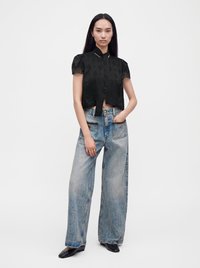 Jeune femme debout portant une blouse noire à manches courtes avec bordure en dentelle, un jean large bleu clair, des chaussures noires et des bagues en or.
