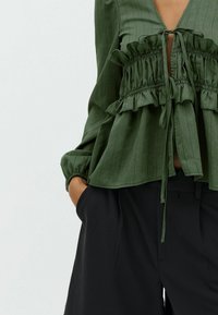 Green blouse made of textured fabric, featuring a deep V-neck, ruffled waistband, and long sleeves with gathered cuffs. Paired with black pants.