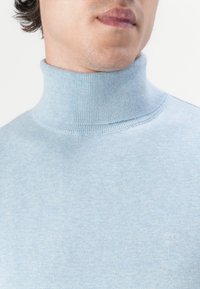 Suéter de cuello alto de color azul claro, fabricado en suave tejido de punto. Presenta un cuello acanalado y un sutil detalle de logo en el pecho.