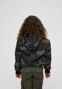 Veste camouflée à capuche, avec un mélange de verts foncés, de gris et de noir. Poignets côtelés et matériau léger.